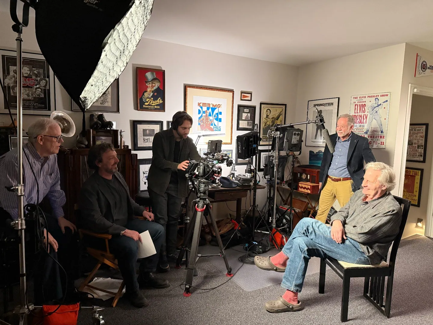 The crew films an interview in a room lined with framed posters, the camera operator at center.