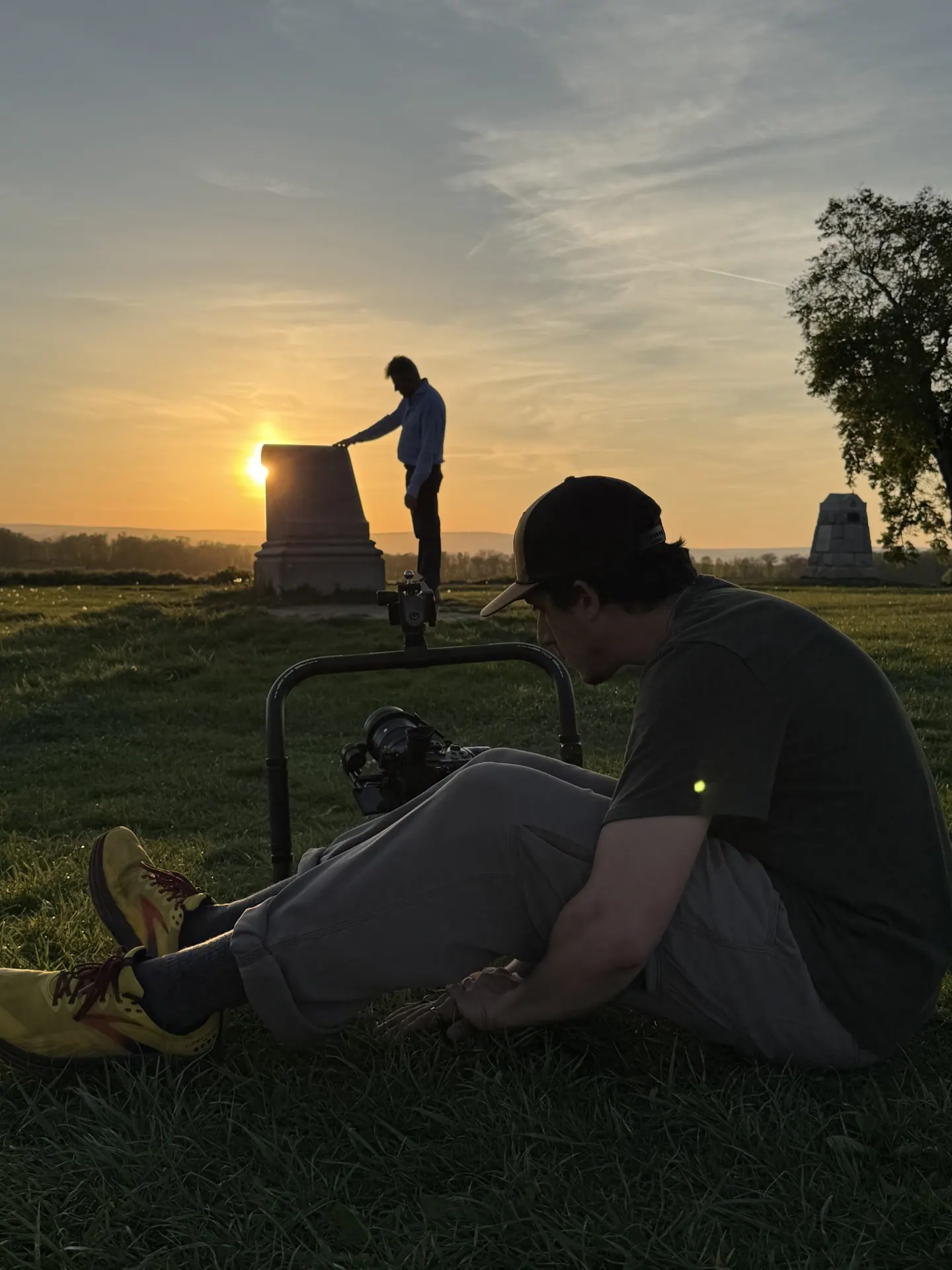 Camera operator seated in the grass filming a figure at a monument at sunset.