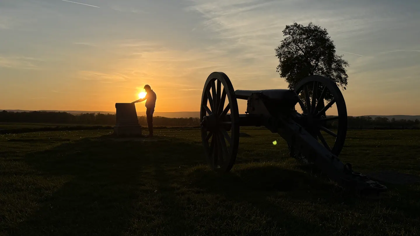Silhouette at sunset beside a monument and a cannon on the battlefield.