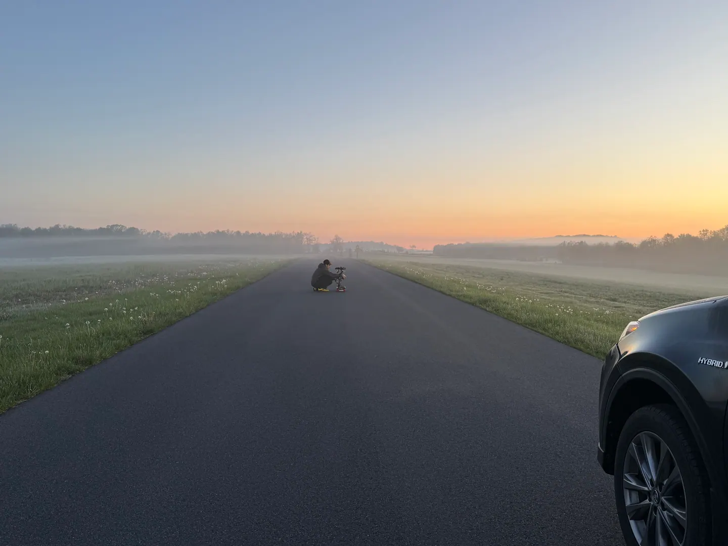 A camera operator crouches on a misty road at dawn.