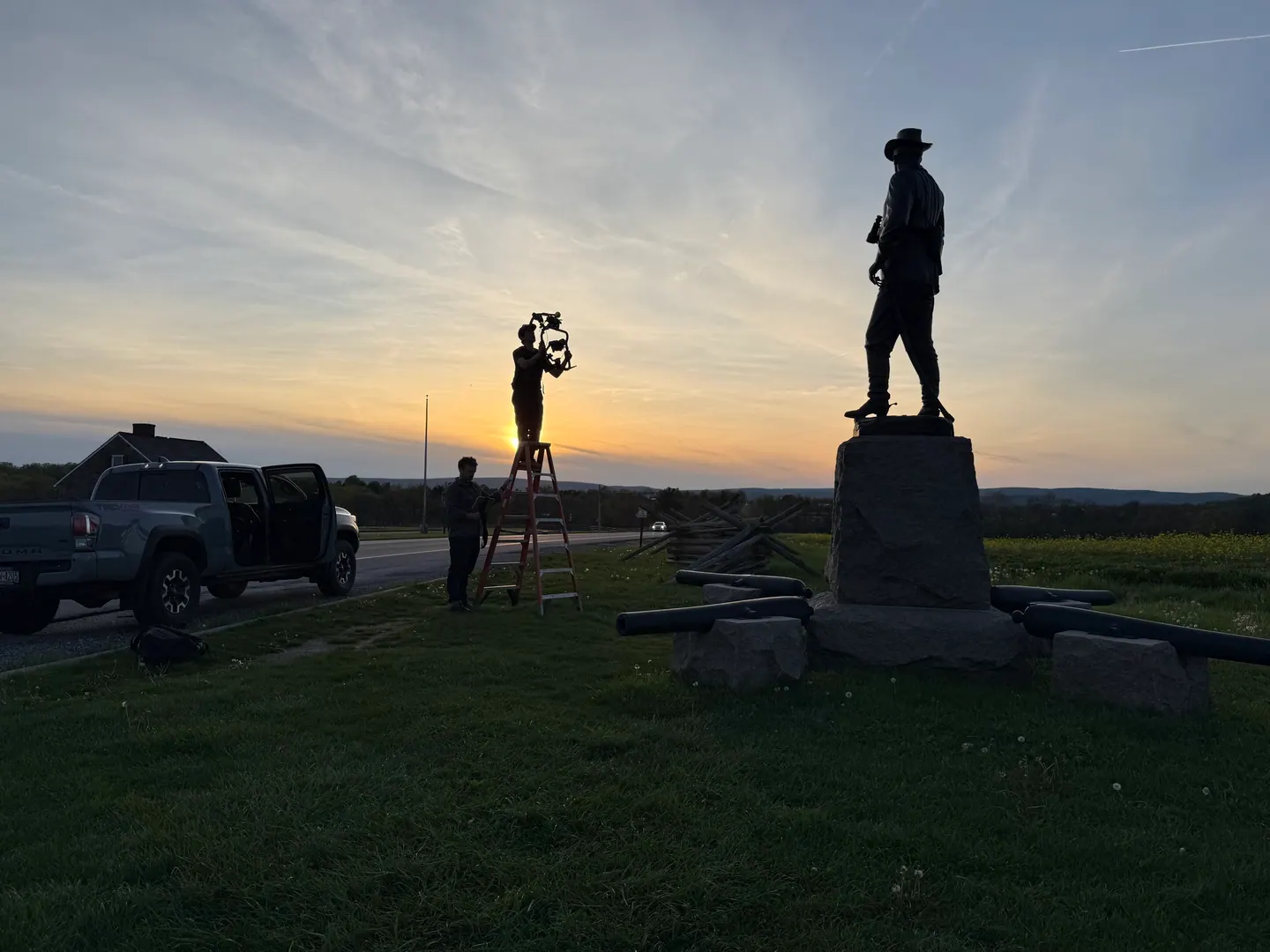 Crew on a ladder filming beside a statue at sunset.