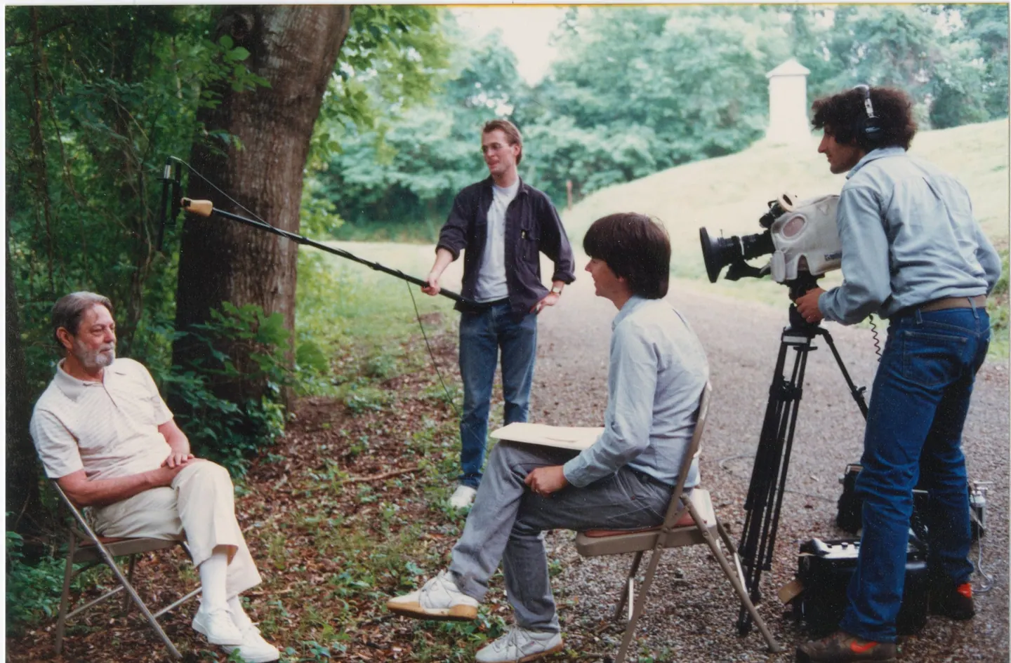 Ken Burns and his crew interview a seated man outdoors beside a tree, with a boom mic and a film camera on a tripod.