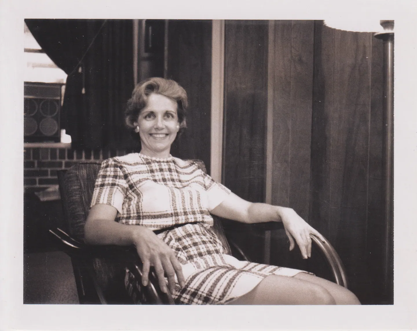 Helen Shaara smiling in a plaid dress, seated in a wooden armchair before wood-panelled walls, 1970s.