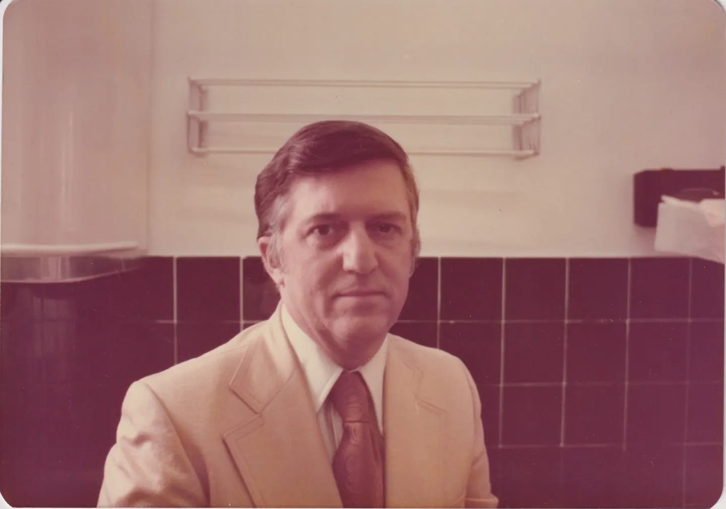 Michael Shaara in a cream suit and patterned tie, seated before a tiled wall, 1977.